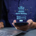 A person holds a smartphone in one hand, displaying an "Online Banking" login interface in holographic style. The other hand types on a laptop keyboard, connecting seamlessly to digital lending platforms. The concept of digital banking security is highlighted. | COYYN