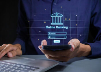 A person holds a smartphone in one hand, displaying an "Online Banking" login interface in holographic style. The other hand types on a laptop keyboard, connecting seamlessly to digital lending platforms. The concept of digital banking security is highlighted. | COYYN