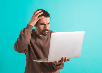 A man in a brown sweater stands against a turquoise background, clutching a laptop in one hand and scratching his head with the other. His face shows confusion or frustration, perhaps pondering how to resolve QuickBooks Error 80029c4a. | COYYN