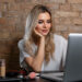 A woman with long blonde hair sits at a desk, chin resting on her hand, focused on a laptop running a desktop video editor. Surrounded by makeup products and illuminated by a ring light, she works intently against the rustic charm of a brick wall backdrop. | COYYN