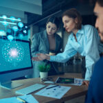 In a modern office, three colleagues focus intently on a computer screen showing a brain graphic labeled "AI," enlivened by digital icons above it. They discuss how AI is helping businesses enhance their designs. A tablet, papers, and a plant add to the lively workspace. | COYYN