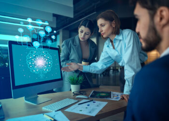 In a modern office, three colleagues focus intently on a computer screen showing a brain graphic labeled "AI," enlivened by digital icons above it. They discuss how AI is helping businesses enhance their designs. A tablet, papers, and a plant add to the lively workspace. | COYYN