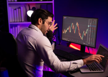 A man in a white shirt sits at a desk with a computer and laptop, analyzing a BYDFi vs MEXC graph on the monitor. He speaks into a microphone, surrounded by bookshelves and ambient purple lighting. | COYYN