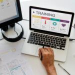 A person is using a laptop that displays an "Employee Training" progress chart, ranging from beginner to expert. Various design tools and sketches are on the desk, alongside a monitor, a notepad, and a plant. The setting suggests a graphic design workspace. | COYYN