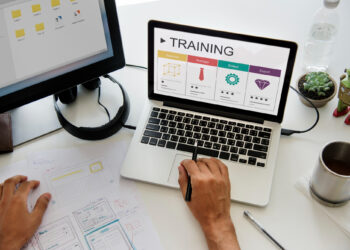 A person is using a laptop that displays an "Employee Training" progress chart, ranging from beginner to expert. Various design tools and sketches are on the desk, alongside a monitor, a notepad, and a plant. The setting suggests a graphic design workspace. | COYYN