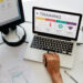 A person is using a laptop that displays an "Employee Training" progress chart, ranging from beginner to expert. Various design tools and sketches are on the desk, alongside a monitor, a notepad, and a plant. The setting suggests a graphic design workspace. | COYYN