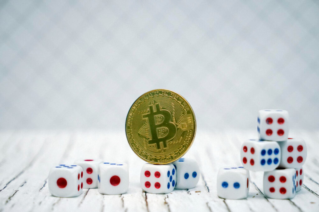 A gold Bitcoin coin stands upright on a wooden surface, surrounded by stacks of Baccarat dice with blue and red dots. The dice are arranged in ascending and descending stacks, creating an intriguing blend of finance and chance against a softly blurred background. | COYYN