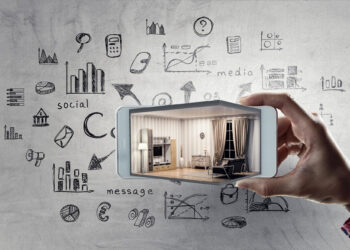 A hand holds a smartphone showing a modern living room. The background features sketches of graphs, charts, and icons related to social media, business, and Digital Marketing Strategy for Your Real Estate Business on a gray surface. | COYYN