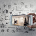 A hand holds a smartphone showing a modern living room. The background features sketches of graphs, charts, and icons related to social media, business, and Digital Marketing Strategy for Your Real Estate Business on a gray surface. | COYYN
