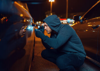 A person in a hoodie seems to be attempting car theft under dim streetlights in a parking lot, using a tool to unlock the door. The scene is dimly lit with reflections dancing on the car’s surface, echoing tales of caution and theft prevention. | COYYN