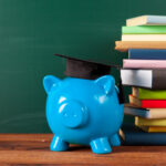 A blue piggy bank wearing a graduation cap sits on a wooden surface next to a stack of books, with a chalkboard in the background. This scene subtly underscores the importance of financial literacy in saving for educational pursuits. | COYYN