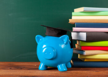 A blue piggy bank wearing a graduation cap sits on a wooden surface next to a stack of books, with a chalkboard in the background. This scene subtly underscores the importance of financial literacy in saving for educational pursuits. | COYYN