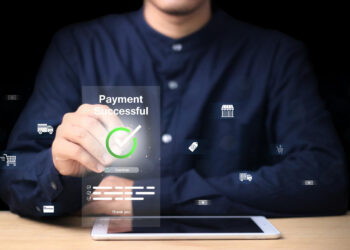 A person in a dark shirt interacts with a virtual screen showing “Payment Successful” and a green checkmark, highlighting Coinme B2B Payment Systems amid digital icons and a tablet on the wooden desk in front of them. | COYYN