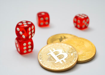 Three red dice and two stacked gold coins with Bitcoin symbols on a white background evoke a sense of risk, much like playing Bitcoin Baccarat, where cryptocurrency meets the thrill of gambling. | COYYN