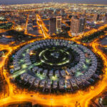 Aerial view of the illuminated Jumeirah Village Circle in new Dubai at night. The circular residential area, surrounded by roads, showcases a striking pattern of buildings contrasted against the cityscape backdrop. | COYYN