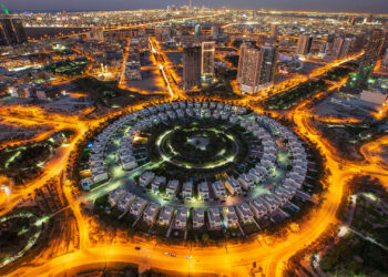 Aerial view of the illuminated Jumeirah Village Circle in new Dubai at night. The circular residential area, surrounded by roads, showcases a striking pattern of buildings contrasted against the cityscape backdrop. | COYYN