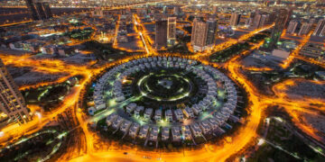 Aerial view of the illuminated Jumeirah Village Circle in new Dubai at night. The circular residential area, surrounded by roads, showcases a striking pattern of buildings contrasted against the cityscape backdrop. | COYYN