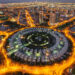 Aerial view of the illuminated Jumeirah Village Circle in new Dubai at night. The circular residential area, surrounded by roads, showcases a striking pattern of buildings contrasted against the cityscape backdrop. | COYYN