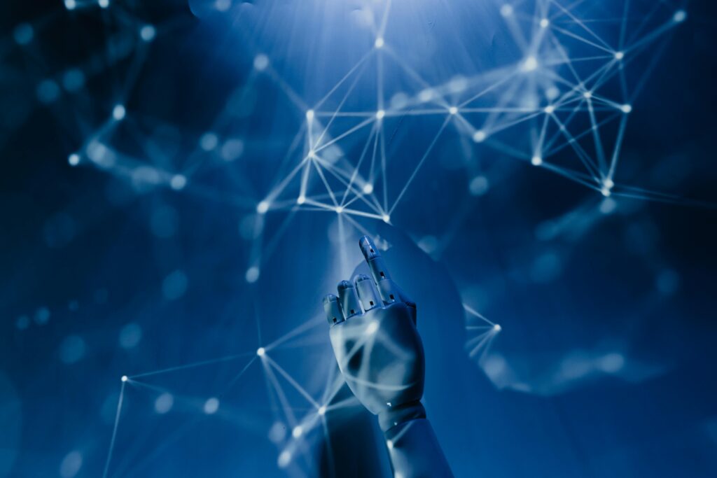 A robotic hand reaches towards a digital network of glowing, interconnected nodes and lines on a blue background, symbolizing advanced technology and artificial intelligence. | COYYN