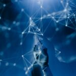 A robotic hand reaches towards a digital network of glowing, interconnected nodes and lines on a blue background, symbolizing advanced technology and artificial intelligence. | COYYN