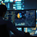 A person sits at a desk in front of multiple screens displaying charts, graphs, and data tracking for crypto enthusiasts, with a city skyline and bright lights visible through the window at night. | COYYN