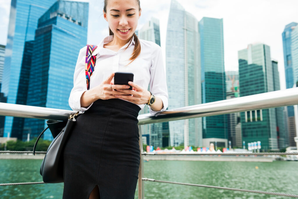 A woman in business attire stands by a railing near the water in Hong Kong, smiling as she uses her phone. Tall modern skyscrapers rise in the background under a bright sky. | COYYN