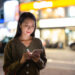 A woman stands outdoors at night in Hong Kong, looking down and smiling at her smartphone, with brightly lit shops and blurred city lights glowing in the background. | COYYN