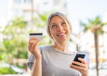 A smiling woman outdoors demonstrates credit card use, holding a card in one hand and a smartphone in the other, with palm trees and buildings softly blurred in the background. | COYYN