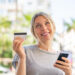 A smiling woman outdoors demonstrates credit card use, holding a card in one hand and a smartphone in the other, with palm trees and buildings softly blurred in the background. | COYYN