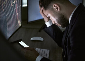 A man in a suit sits at a desk looking stressed, holding his head and checking his phone. Stock charts show falling prices on the monitors, suggesting he may be affected by digital investment scams. | COYYN