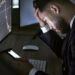A man in a suit sits at a desk looking stressed, holding his head and checking his phone. Stock charts show falling prices on the monitors, suggesting he may be affected by digital investment scams. | COYYN