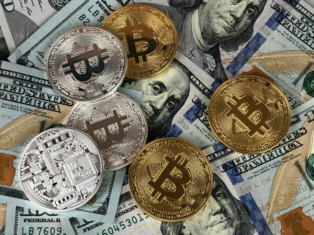 A mix of gold and silver Bitcoin coins placed on top of scattered U.S. one hundred dollar bills, highlighting the dynamic relationship between physical currency and the fluctuating Bitcoin Price USD. | COYYN