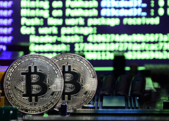 Two physical Bitcoin coins stand upright on a computer motherboard, with blurred green and blue computer code in the background, symbolizing cryptocurrency mining and the role of crypto exchanges in digital asset transactions. | COYYN