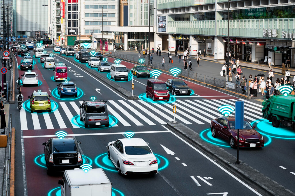 A busy city street with many cars at a crosswalk, each vehicle surrounded by glowing blue wireless symbols, illustrating how connected technology in smart vehicles helps prevent accidents. Pedestrians stand on the sidewalk. | COYYN