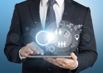 A man in a suit uses a tablet displaying holographic graphs, icons of people, gears, and location markers, symbolizing business analytics and digital technology. | COYYN