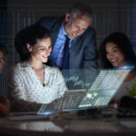 Three people sit at a table, smiling and looking at a futuristic transparent screen displaying digital charts and data, with one standing behind them, in an office setting. | COYYN