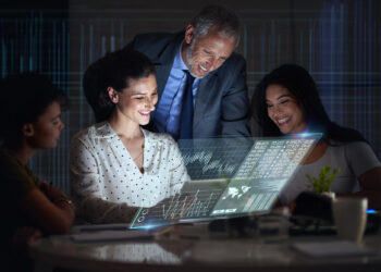 Three people sit at a table, smiling and looking at a futuristic transparent screen displaying digital charts and data, with one standing behind them, in an office setting. | COYYN