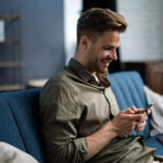 A smiling man sits on a blue sofa, enjoying his smartphone and the thrill of an online casino experience. Casually dressed in a green shirt, he relaxes in a cozy living room with shelves and a wooden cabinet in the background. | COYYN