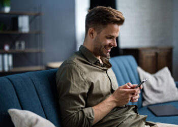 A smiling man sits on a blue sofa, enjoying his smartphone and the thrill of an online casino experience. Casually dressed in a green shirt, he relaxes in a cozy living room with shelves and a wooden cabinet in the background. | COYYN