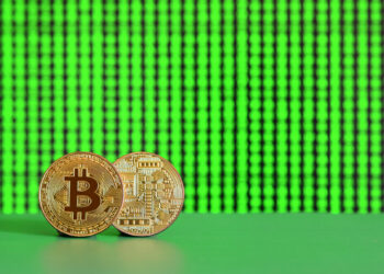 Two physical Bitcoin coins stand upright on a green surface with a bright green digital matrix background, symbolizing cryptocurrency and digital finance. | COYYN