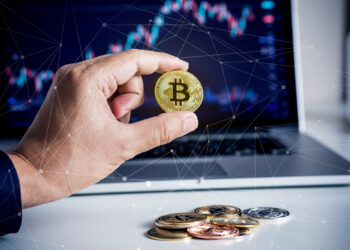 A hand holds a gold Bitcoin coin in front of a laptop displaying a financial chart, with several other cryptocurrency coins scattered on the desk. Digital connection lines overlay the image, highlighting blockchain and CEX.IO: A Trusted and User-Friendly Crypto Exchange. | COYYN
