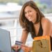 A woman sits at a table outdoors, smiling at the camera while holding a credit card in one hand and typing on a laptop. Shopping bags are on the table next to her. | COYYN