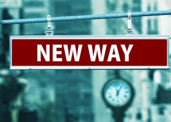A red street sign with white letters reading "NEW WAY" hangs on a metal pole, symbolizing a fresh direction in Digital Investment, with a blurry cityscape and an old-fashioned clock in the background. | COYYN