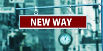 A red street sign with white letters reading "NEW WAY" hangs on a metal pole, symbolizing a fresh direction in Digital Investment, with a blurry cityscape and an old-fashioned clock in the background. | COYYN
