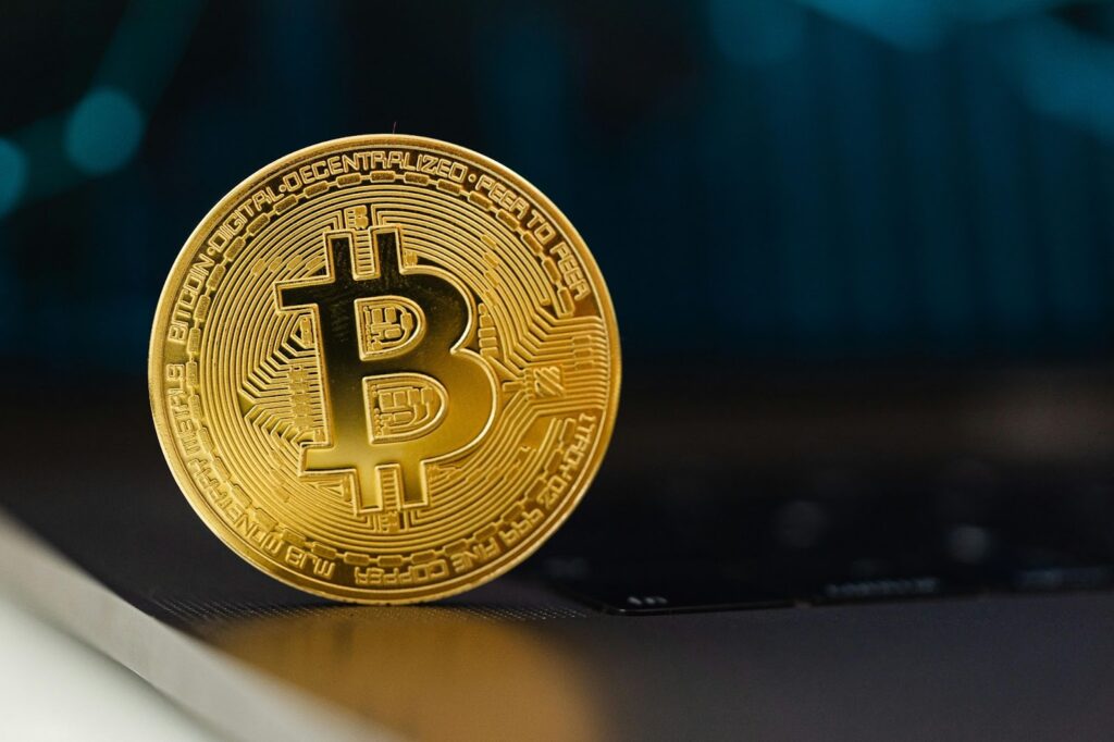 A gold-colored physical Bitcoin coin with the Bitcoin symbol stands upright on the edge of a laptop keyboard, evoking the thrill of gambling against a blurred dark blue background. | COYYN
