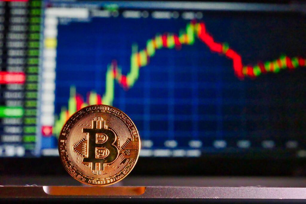 A gold Bitcoin coin stands in front of a blurred screen showing a cryptocurrency price chart with red and green candlesticks, symbolizing market fluctuations and the importance of securing assets in a trusted crypto wallet. | COYYN
