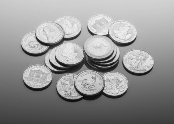 A scattered pile of various silver bullion coins featuring different designs—animals, people, and symbols—rests on a smooth grey surface. | COYYN