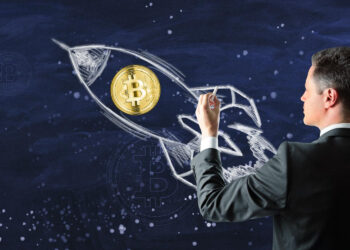 A man in a suit draws a white chalk rocket on a dark blue chalkboard, with a gold Bitcoin symbol placed inside the rocket, symbolizing cryptocurrency growth. | COYYN