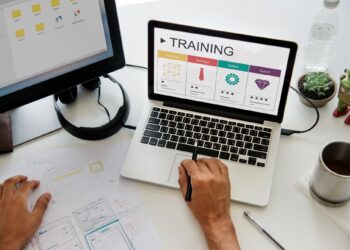 Person using a laptop displaying a training program, with design sketches and office items on the desk.