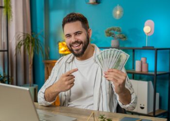 Smiling man at a desk points to a fan of dollar bills, sitting in front of a laptop in a modern room.